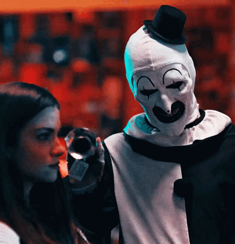 A Woman Stands Next To A Clown Wearing A Mask And Top Hat GIF