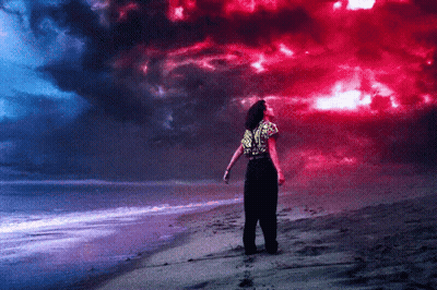 A Woman Stands On A Beach Looking Up At A Red And Blue Sky GIF