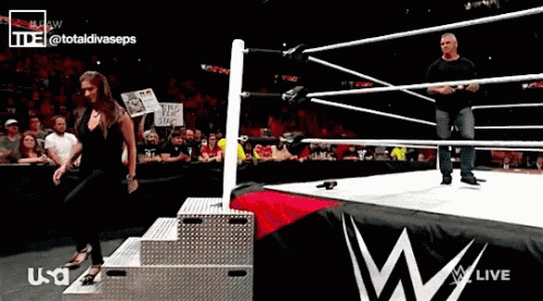 A Woman Stands On A Podium In A Wrestling Ring While A Man Stands In The Ring GIF