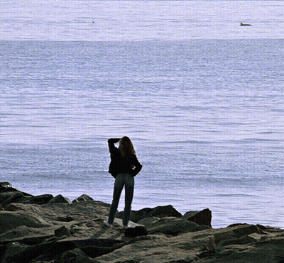 A Woman Stands On A Rocky Shore Looking Out Over The Ocean GIF