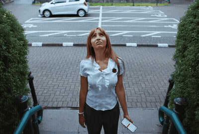 A Woman Stands On A Set Of Stairs Looking Up At A Parking Lot With A White Car Parked In It GIF