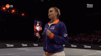 A Woman Stands On A Tennis Court Wearing A Blue And Orange Jacket GIF