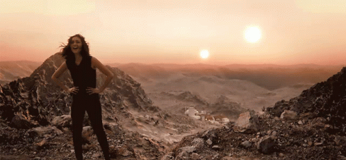 A Woman Stands On Top Of A Rocky Mountain With The Sun Behind Her GIF