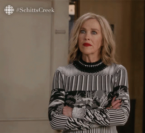 A Woman Stands With Her Arms Crossed In Front Of A Sign That Says Schitts Creek On It GIF