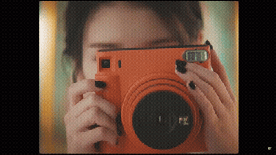 A Woman Taking A Picture With An Orange Camera That Says Fuji GIF