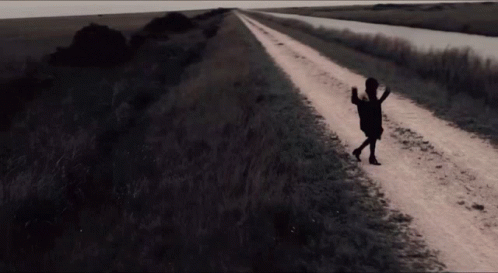 A Woman Walking Down A Dirt Road With A Purse In Her Hand GIF