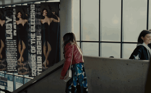 A Woman Walking Down A Set Of Stairs Next To A Poster That Says Your 2020 Bleeding GIF