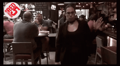 A Woman Walking Through A Crowded Restaurant With A Red R25 Network Logo Behind Her GIF