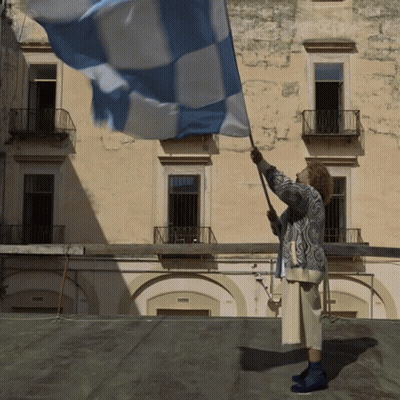 A Woman Waves A Blue And White Flag In Front Of A Building GIF