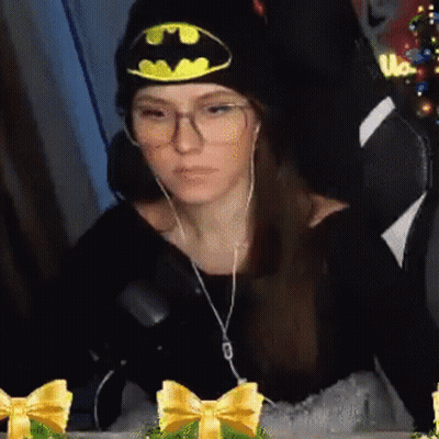 A Woman Wearing A Batman Hat And Glasses Is Sitting In A Chair With Headphones On . GIF