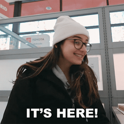 A Woman Wearing A Beanie And Glasses Is Smiling And Says It 'S Here GIF