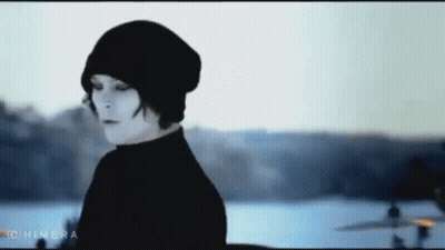 A Woman Wearing A Black Beanie And A Black Turtleneck Is Standing In Front Of A Body Of Water . GIF