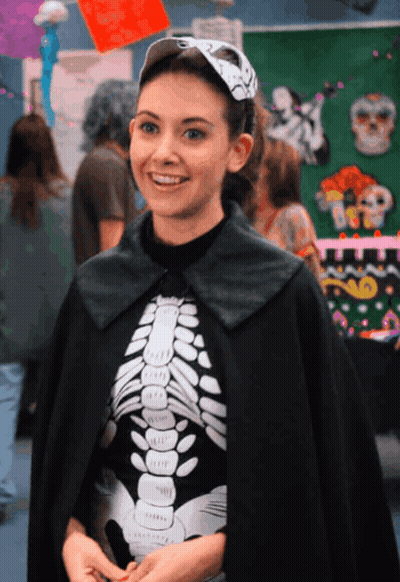 A Woman Wearing A Black Cape And A Skeleton Shirt Smiles GIF