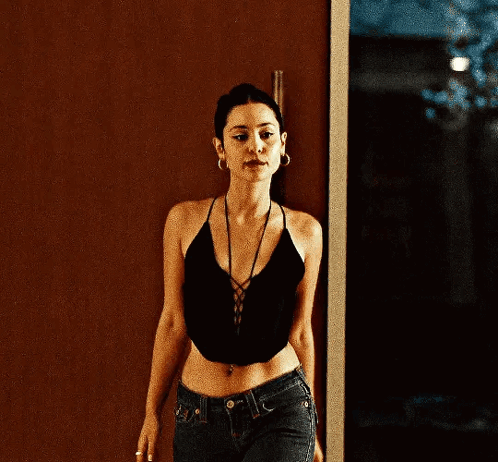 A Woman Wearing A Black Crop Top And Jeans Is Standing In Front Of A Door . GIF
