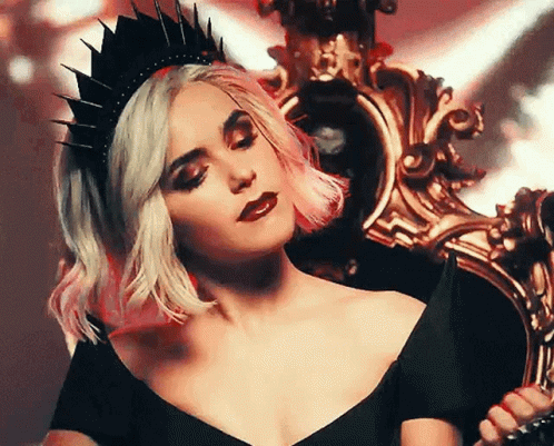 A Woman Wearing A Black Crown And A Black Dress Is Sitting On A Chair . GIF