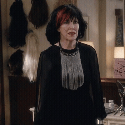 A Woman Wearing A Black Dress And A Silver Necklace Stands In Front Of A Mirror GIF