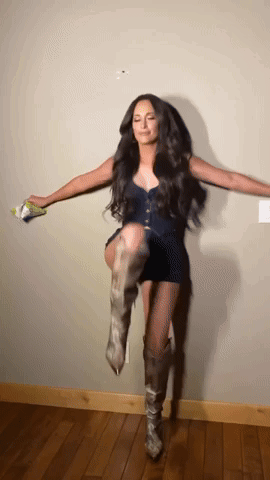 A Woman Wearing A Black Dress And Cowboy Boots Is Standing On One Leg GIF