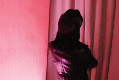 A Woman Wearing A Black Hat And A Leather Jacket Is Standing In Front Of A Pink Curtain GIF