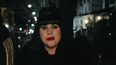A Woman Wearing A Black Hat And Red Lipstick Is Standing On A City Street At Night . GIF