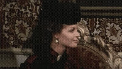 A Woman Wearing A Black Hat Is Sitting On A Bed GIF