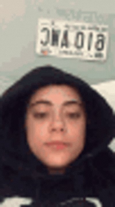 A Woman Wearing A Black Hoodie Is Making A Funny Face In Front Of A License Plate . GIF