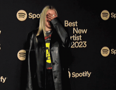A Woman Wearing A Black Jacket And A Yellow Shirt Stands In Front Of A Sign That Says Best New Artist 2023 GIF