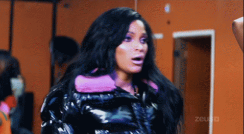 A Woman Wearing A Black Jacket With A Pink Hood Is Standing In Front Of An Orange Wall With The Word Zeus Written On It GIF