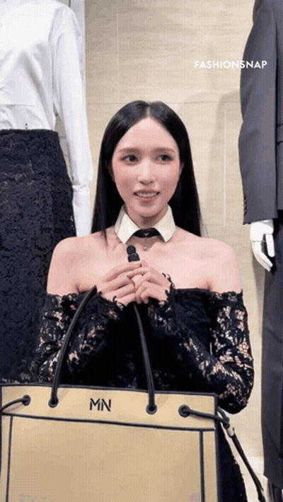 A Woman Wearing A Black Lace Off The Shoulder Top Is Holding A Bag With The Letter Mn On It GIF