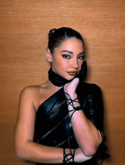 A Woman Wearing A Black Leather Dress And Gloves Is Standing In Front Of A Wooden Wall GIF