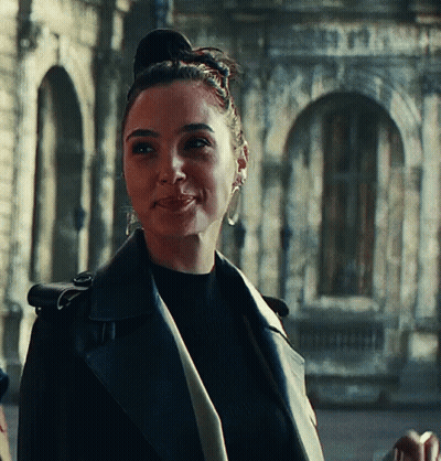 A Woman Wearing A Black Leather Jacket And Earrings Smiles In Front Of A Building GIF
