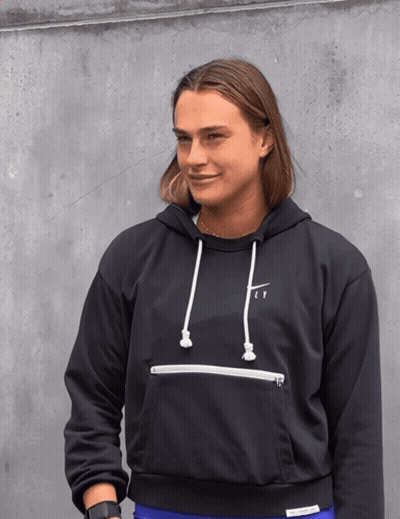 A Woman Wearing A Black Nike Hoodie Stands In Front Of A Grey Wall GIF