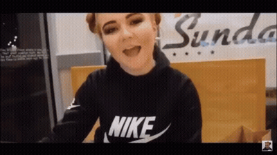 A Woman Wearing A Black Nike Sweatshirt Is Sitting In Front Of A Sign That Says Sunda . GIF