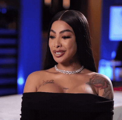 A Woman Wearing A Black Off The Shoulder Top And A Necklace Is Sitting On A Bed . GIF