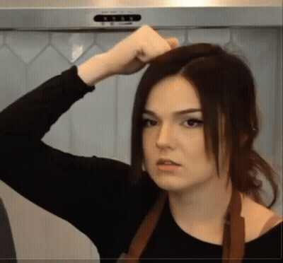 A Woman Wearing A Black Shirt And An Apron Holds Her Hair Up GIF