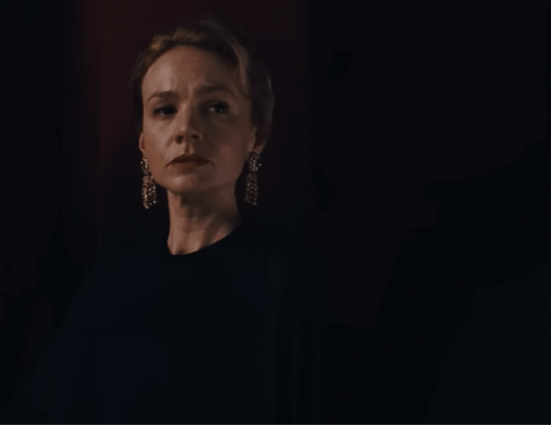A Woman Wearing A Black Shirt And Earrings Looks At The Camera In A Dark Room GIF
