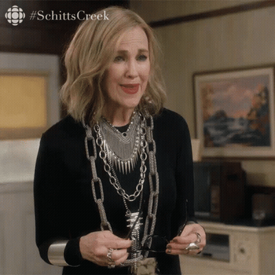A Woman Wearing A Black Shirt And Silver Jewelry Is Holding A Pair Of Glasses With The Hashtag Schitts Creek On The Top GIF