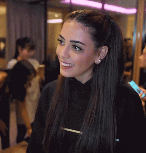 A Woman Wearing A Black Sweater With The Letter T On The Front Smiles GIF