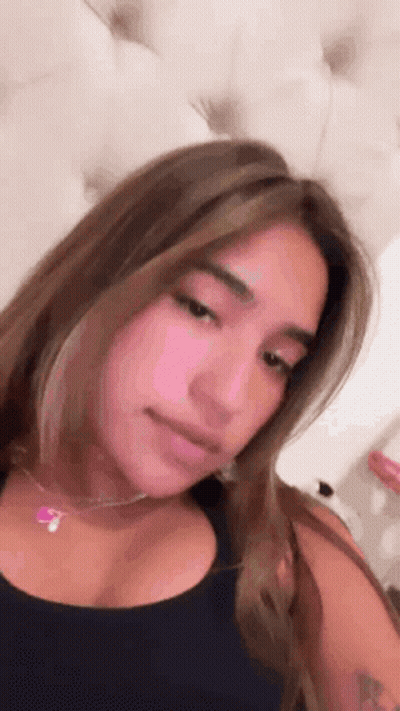 A Woman Wearing A Black Tank Top And A Pink Necklace Is Sitting On A Bed . GIF