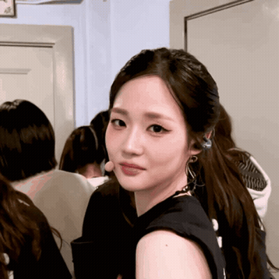 A Woman Wearing A Black Tank Top And Earrings Looks Over Her Shoulder GIF