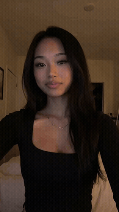 A Woman Wearing A Black Top And A Necklace Is Standing In A Dark Room . GIF