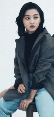 A Woman Wearing A Black Turtleneck And A Grey Jacket Sits On A Stool GIF