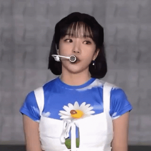 A Woman Wearing A Blue And White Shirt With A Daisy On It Is Holding A Microphone In Her Mouth . GIF