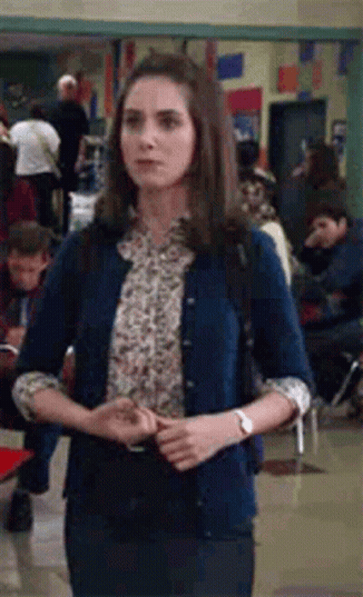 A Woman Wearing A Blue Cardigan And A Floral Shirt Is Standing In A Hallway . GIF