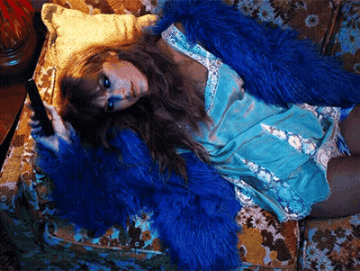 A Woman Wearing A Blue Fur Coat Is Laying On A Couch Holding A Cell Phone . GIF