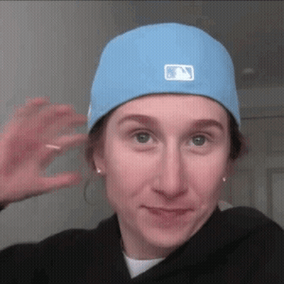 A Woman Wearing A Blue Hat And A Black Hoodie Is Waving At The Camera . GIF