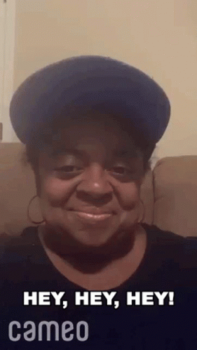 A Woman Wearing A Blue Hat And Earrings Is Sitting On A Couch And Smiling . GIF