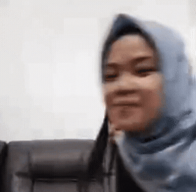 A Woman Wearing A Blue Hijab Is Sitting On A Couch And Smiling . GIF