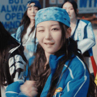 A Woman Wearing A Blue Jacket And A Blue Bandana Is Smiling In Front Of A Sign That Says But Always Open GIF