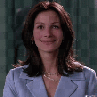 A Woman Wearing A Blue Jacket And A Silver Necklace Smiles For The Camera GIF