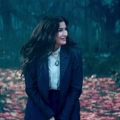 A Woman Wearing A Blue Jacket And White Shirt Is Standing In A Forest . GIF